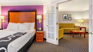 A hotel room with a large bed, a bedside table with a lamp, a yellow armchair, and a small wooden table.