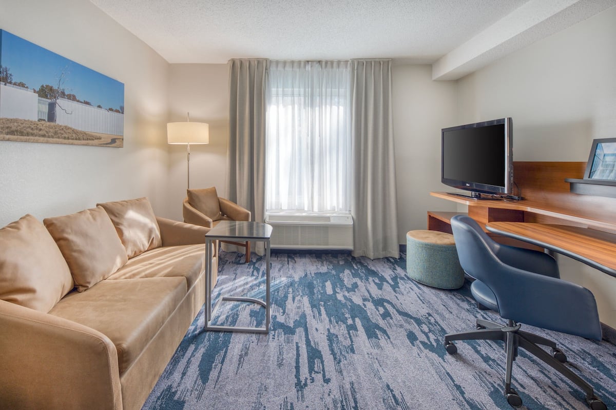 A hotel room with a beige sofa, a small table, a TV, and a desk with a chair.