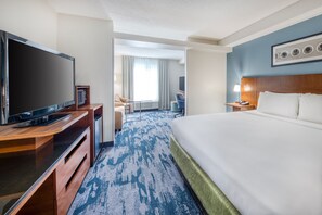 A hotel room with a large bed, a flat-screen TV, a desk with a computer, and a view of the hotel lobby.