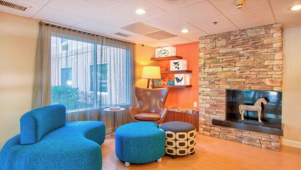 A modern living room with a stone fireplace, blue upholstered furniture, and a view of the outdoors through large windows. - Fairfield Inn By Marriott Raleigh Crabtree (Raleigh)