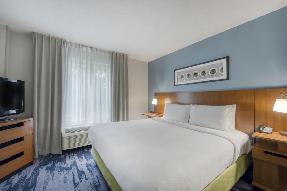 A hotel room with a large bed, a television on a wooden dresser, a window with sheer curtains, and a wall-mounted picture frame.