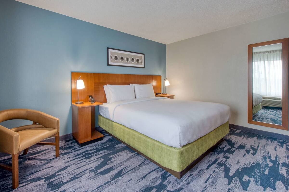 A hotel room with a bed, a chair, a nightstand, and a wall-mounted picture.