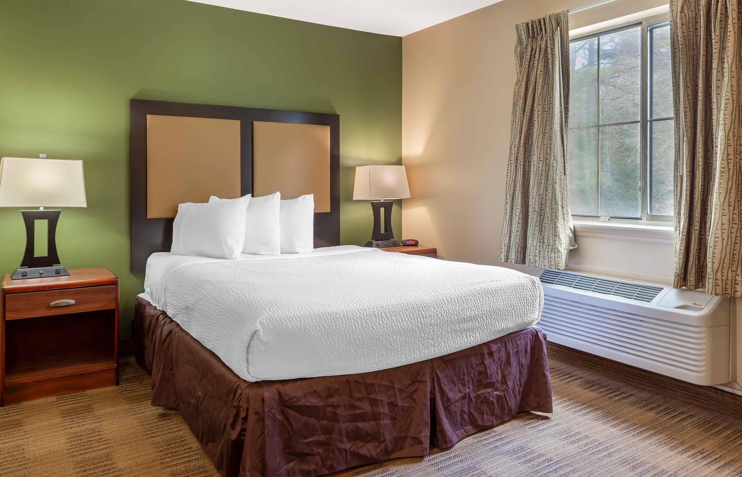 Una habitación de hotel con una cama grande, dos mesitas de noche, una ventana con cortinas y un aire acondicionado de pared.