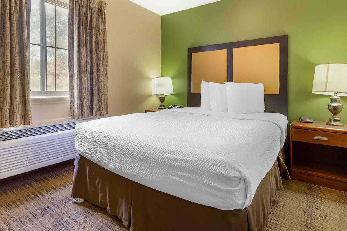 Ein Hotelzimmer mit einem großen Bett, zwei Nachttischen mit Lampen, einem Fenster mit Vorhängen und einer Wand-Klimaanlage.