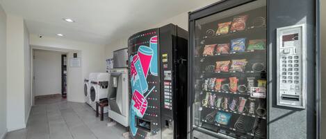 A vending machine with snacks and a coin-operated machine, a washing machine, and a dryer in a hallway.
