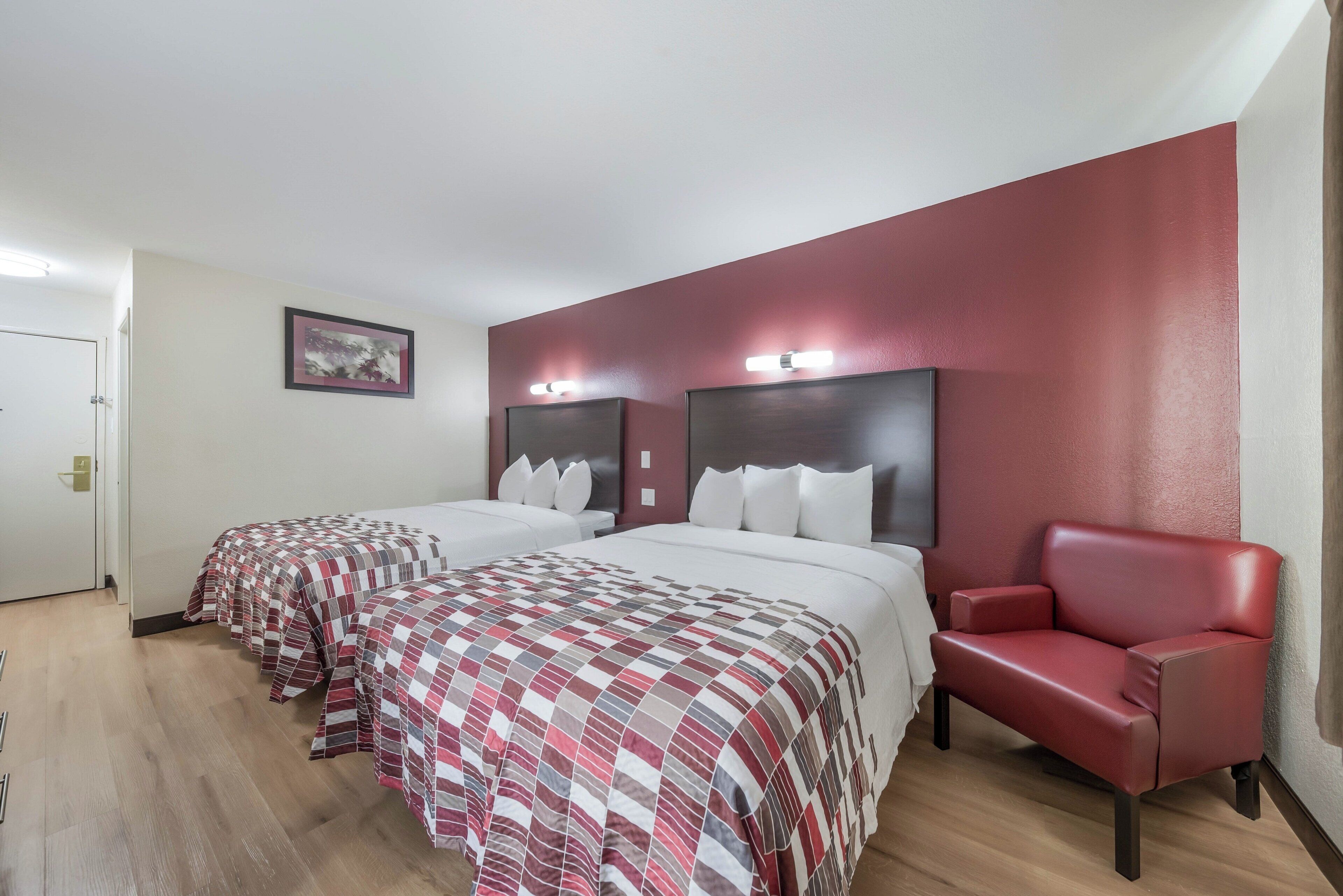A hotel room with two beds, a red leather chair, and a framed picture on the wall.