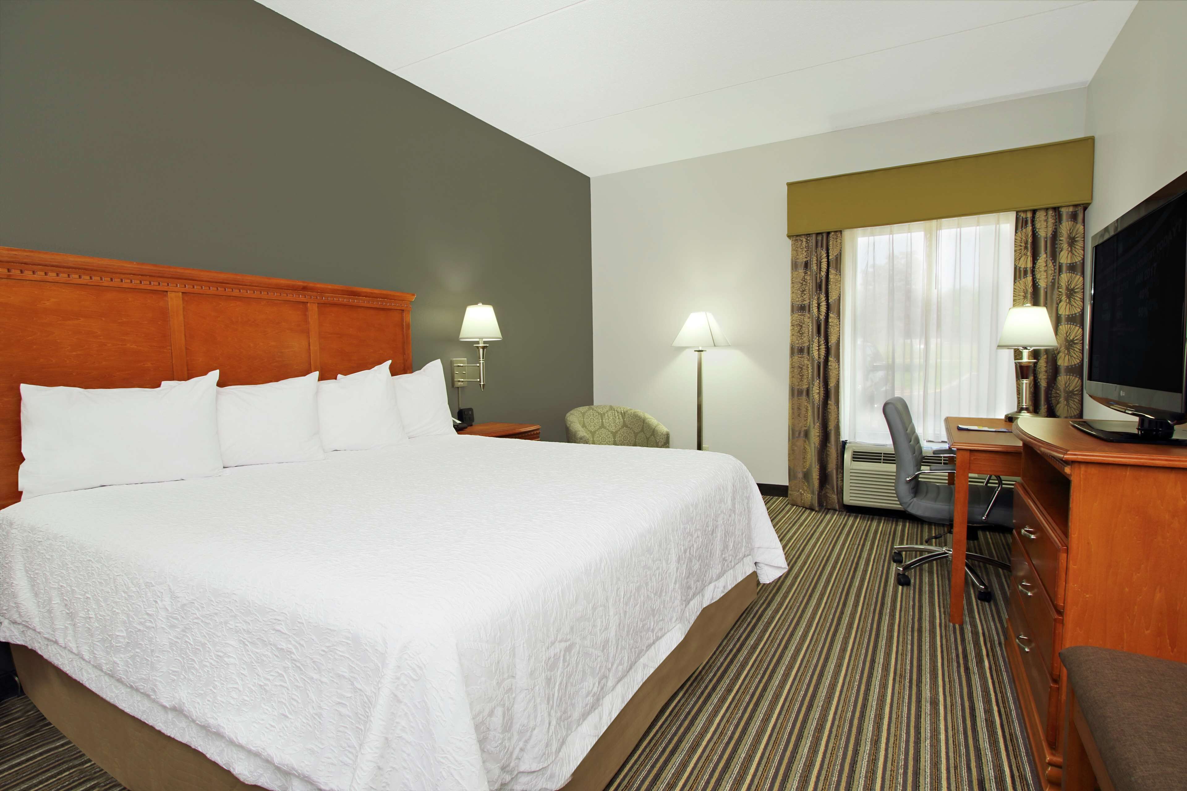 Habitación de hotel con una cama grande, dos mesitas de noche con lámparas, un escritorio con silla, un televisor y una ventana con cortinas.