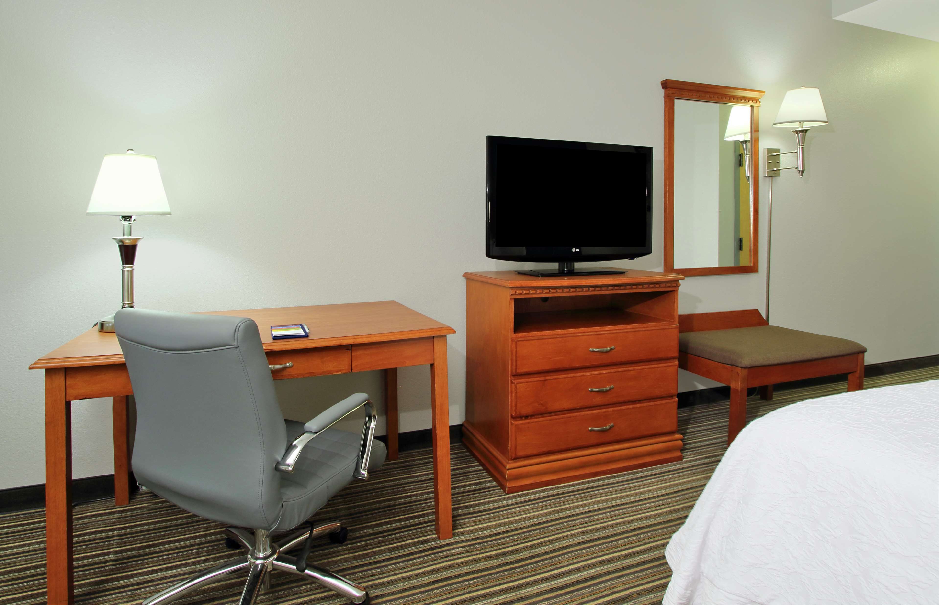 A hotel room with a wooden desk, a television, a mirror, a lamp, a chair, and a bed.