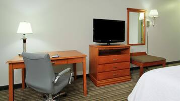 A hotel room with a wooden desk, a television, a mirror, a lamp, a chair, and a bed.