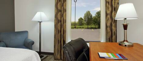 A hotel room with a bed, a desk with a lamp, a chair, and a view of a parking lot through the window.