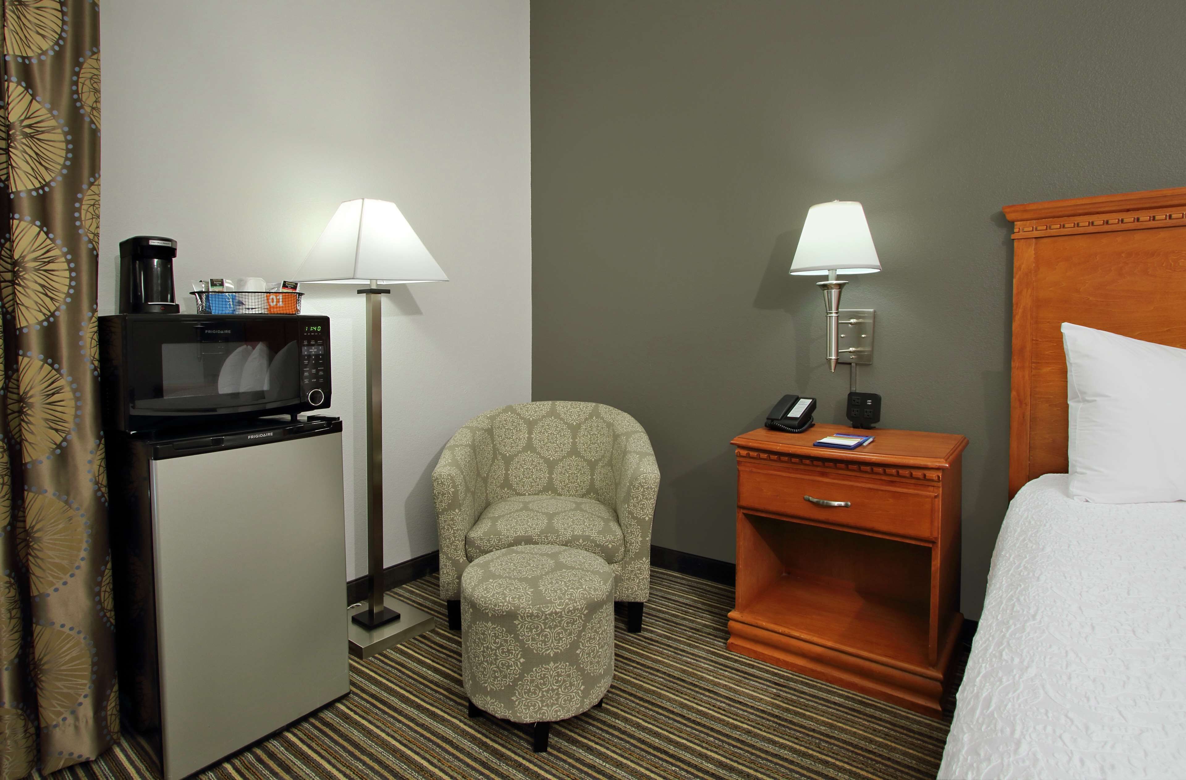 A hotel room with a microwave, a chair, a bedside table with a lamp, and a bed.