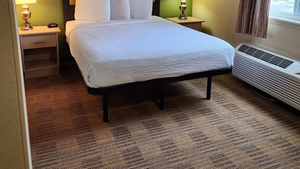 A hotel room with a large bed, two bedside tables with lamps, a window with curtains, and a wall-mounted air conditioner. - Suburban Studios Columbus - Dublin (Dublin)