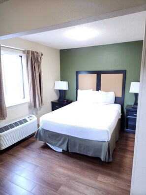 A hotel room with a large bed, two bedside tables with lamps, a window with curtains, and an air conditioning unit. - Suburban Studios Columbus - Dublin (Dublin)