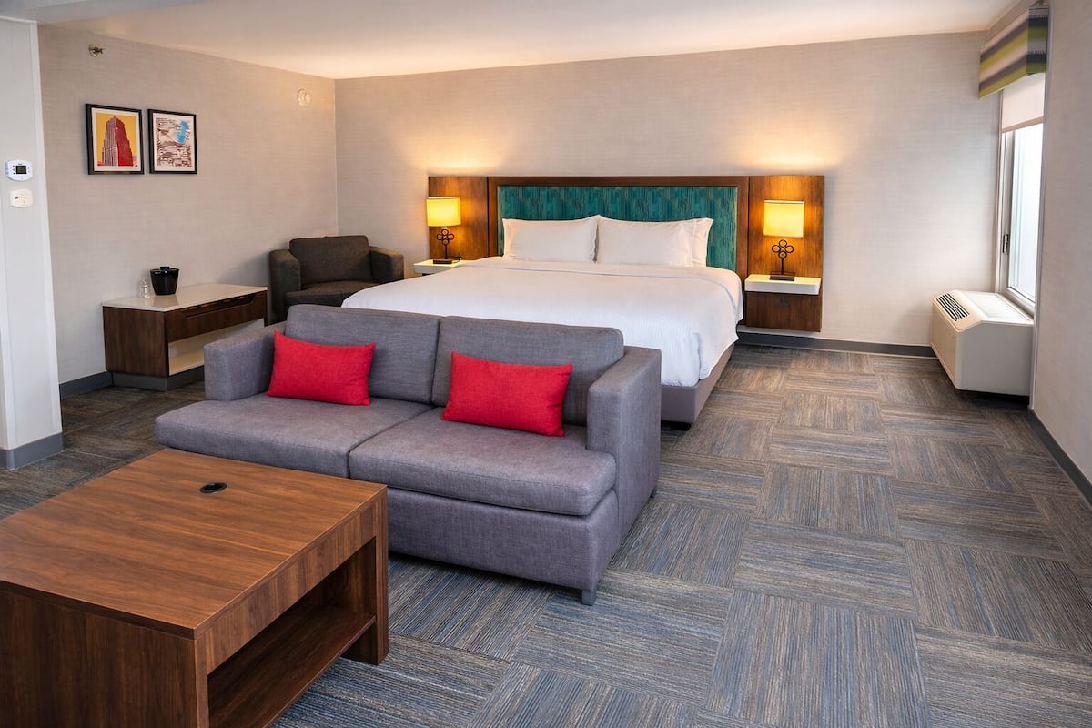 A hotel room with a bed, a grey sofa with red cushions, a wooden coffee table, and two bedside lamps.