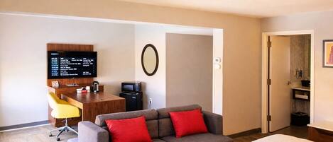 A hotel room with a bed, a sofa with red cushions, a desk with a computer and a coffee maker, and a TV on the wall.