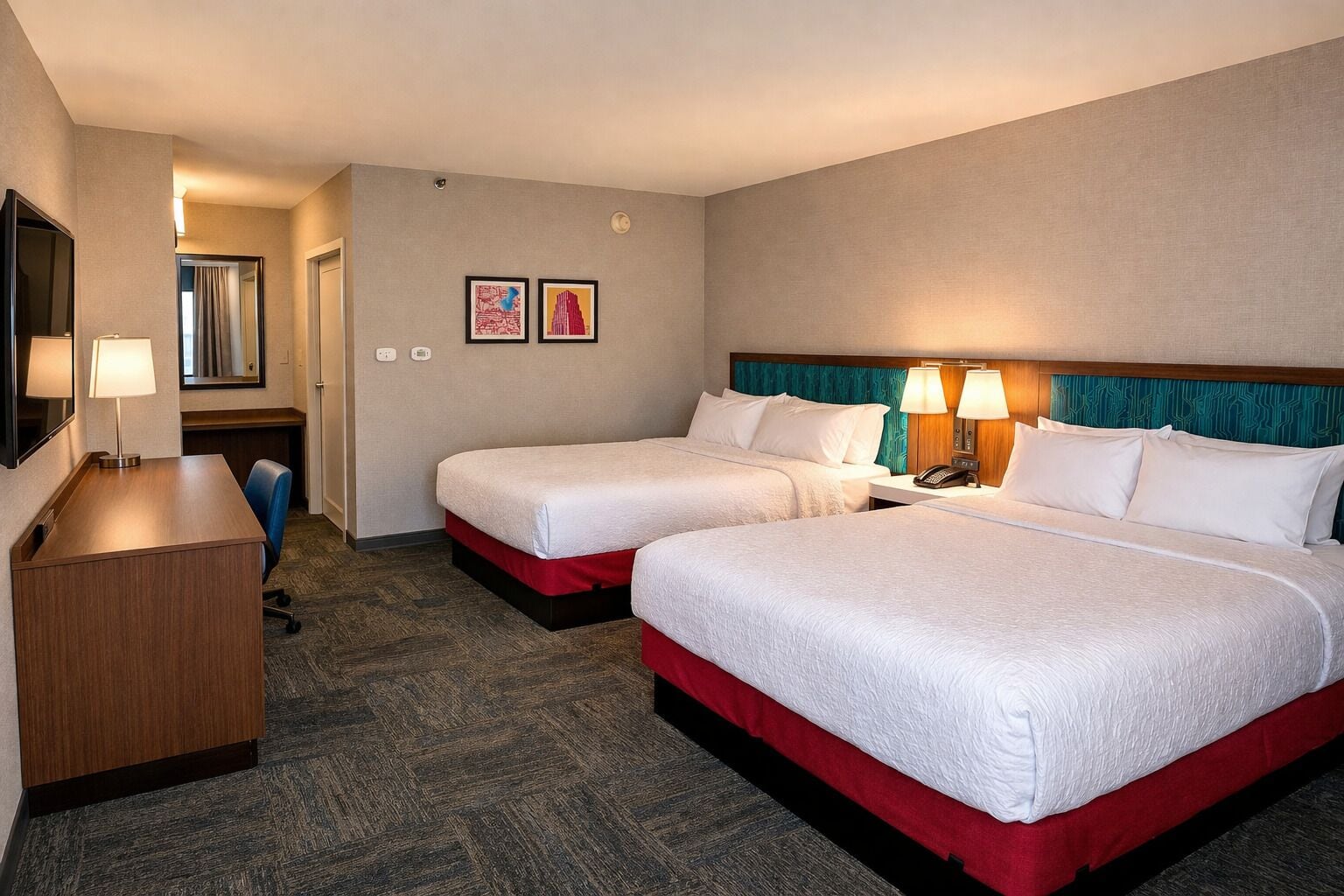 A hotel room with two beds, a desk, a chair, a mirror, and two framed pictures on the wall.