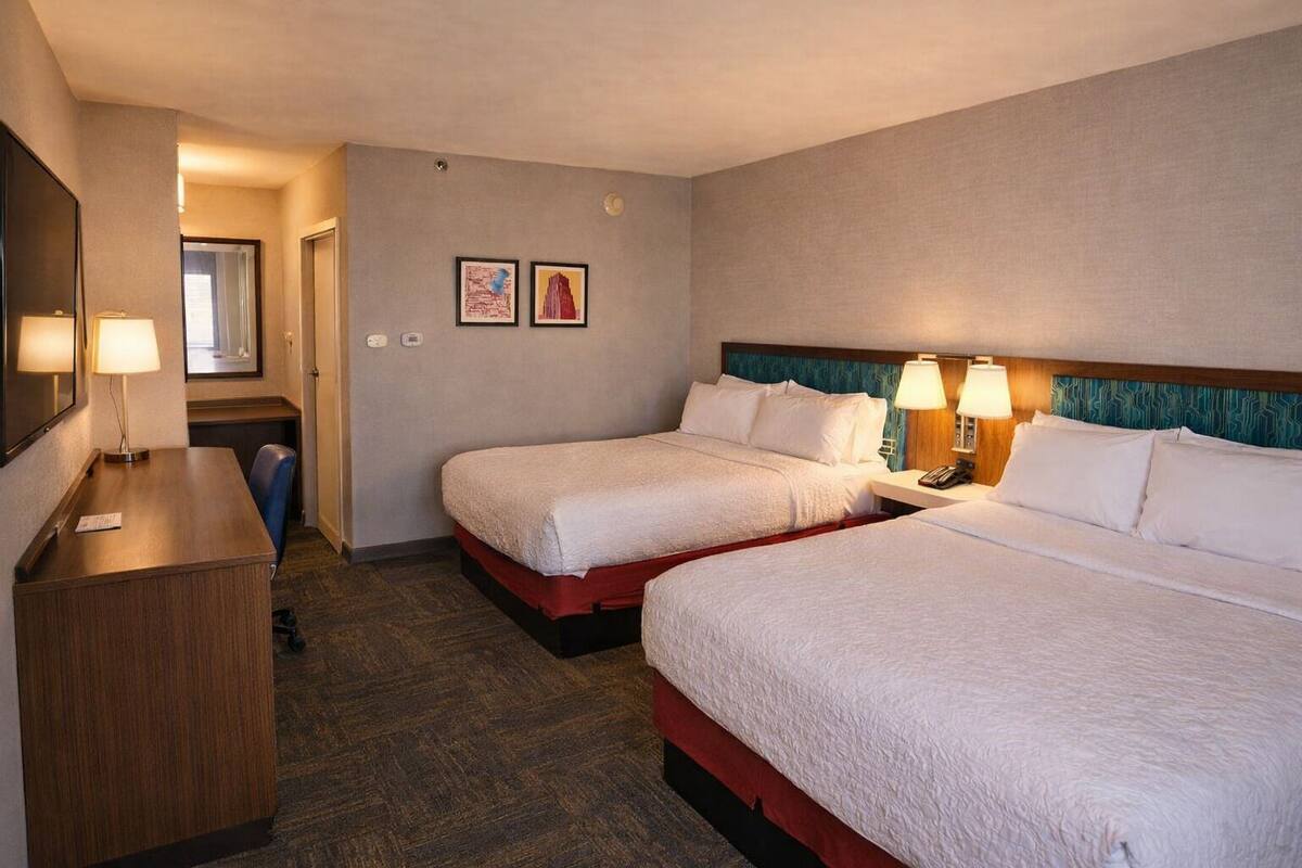 A hotel room with two beds, a desk, a mirror, and two framed pictures on the wall.
