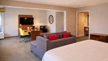 Quarto de hotel com cama, sofá, escrivaninha com televisão, espelho e porta para um banheiro.