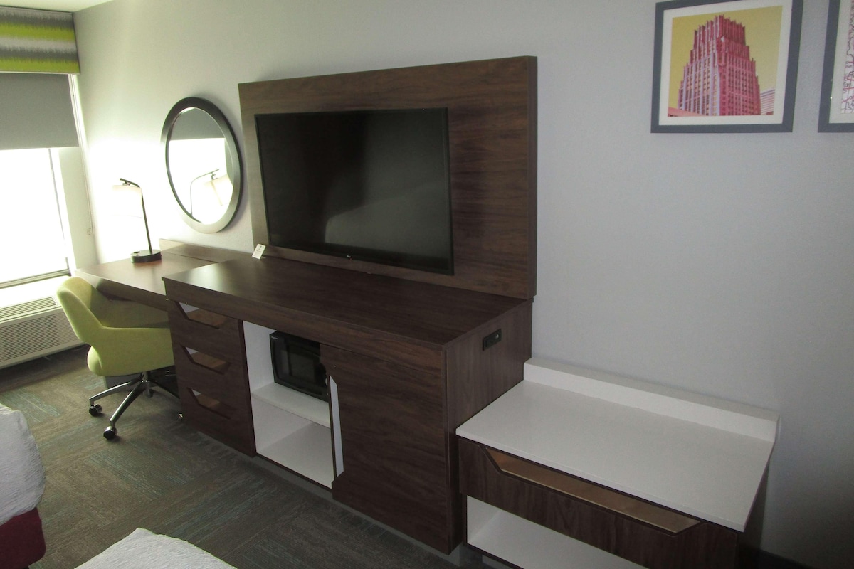 A hotel room with a TV, a desk, a chair, a mirror, and a framed picture on the wall.
