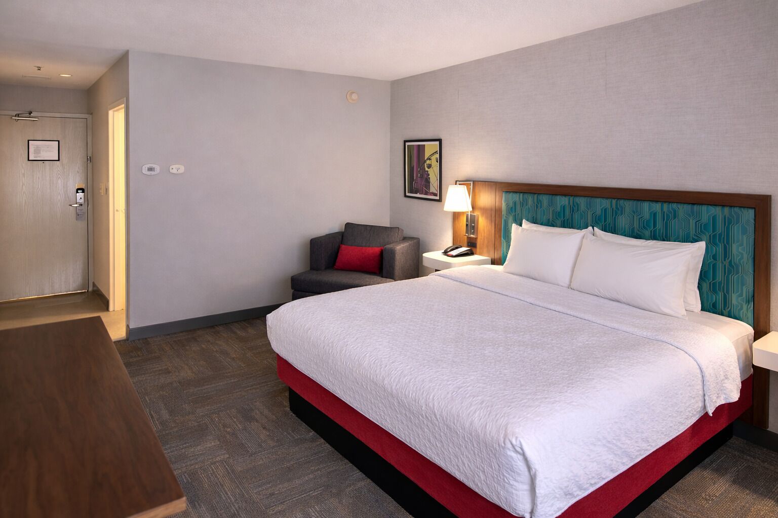 A hotel room with a large bed, a desk, a chair, a nightstand, and a door.