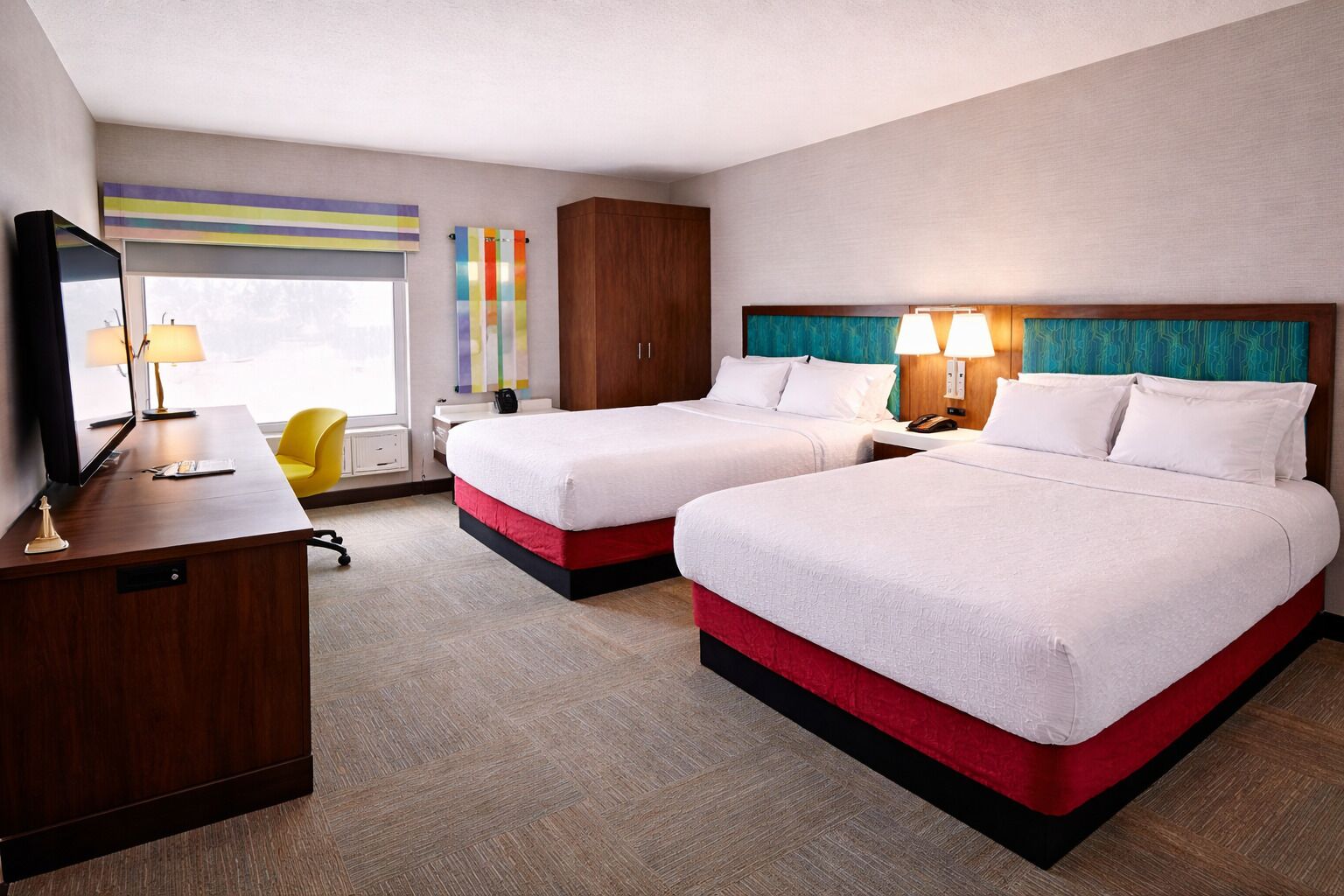 Quarto de hotel com duas camas, uma escrivaninha, uma cadeira, televisão e janela com persianas.