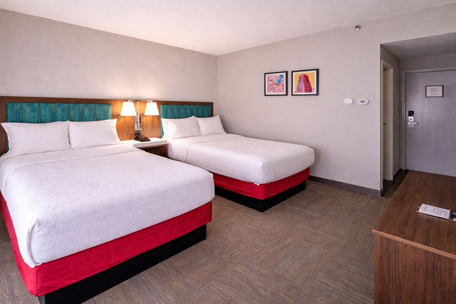 Habitación de hotel con dos camas, una mesita de noche con lámparas, un escritorio y dos obras de arte enmarcadas en la pared.