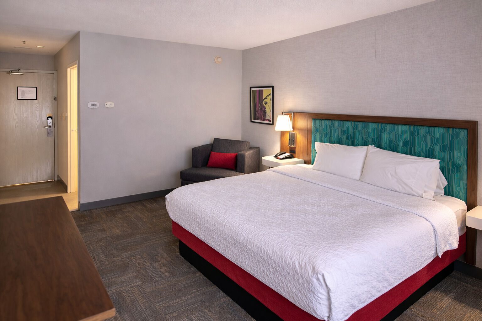 Una habitación de hotel con una cama grande, un escritorio, una silla, una mesita de noche y una puerta.