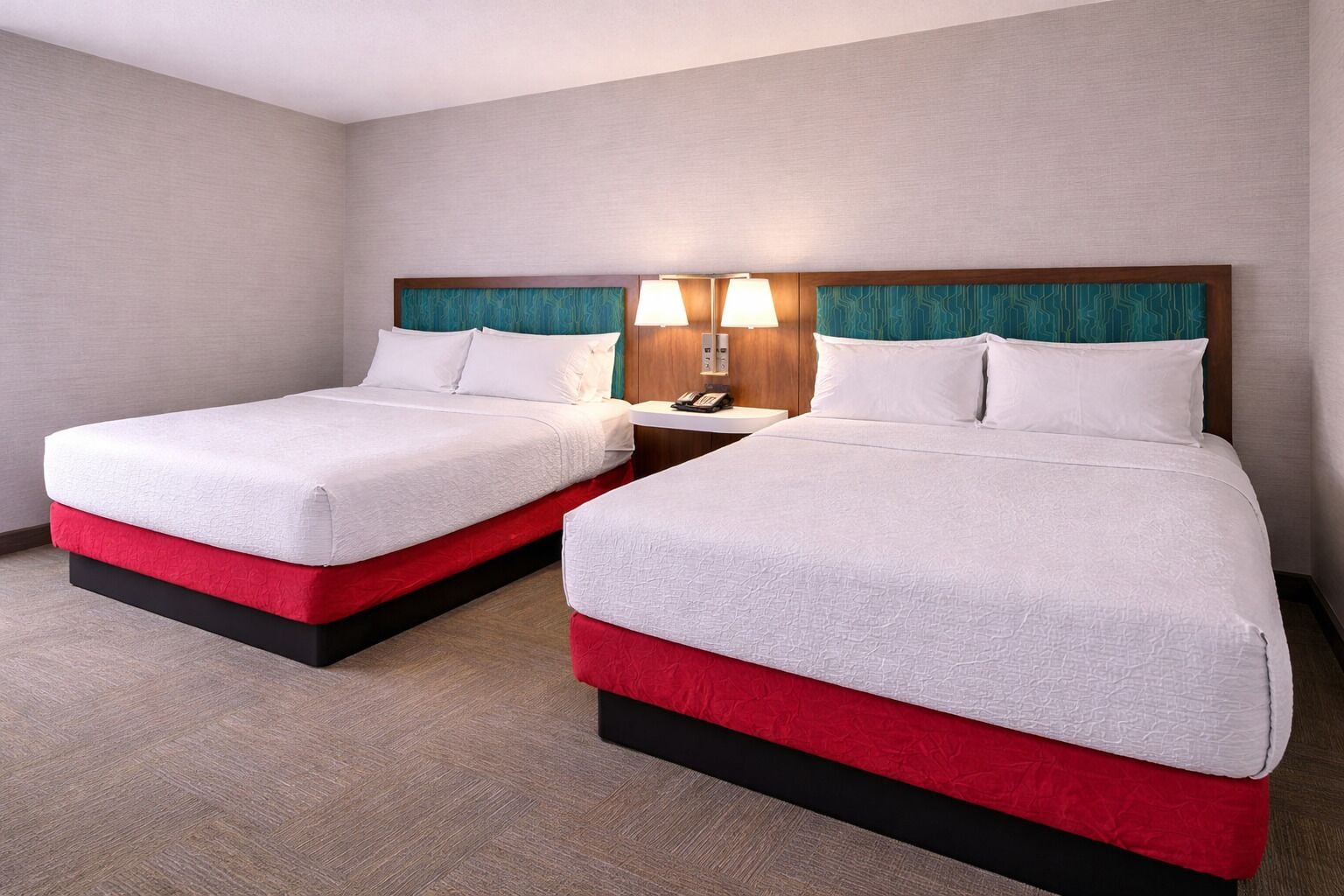 Quarto de hotel com duas camas, mesa de cabeceira com telefone e luminária fixada na parede.