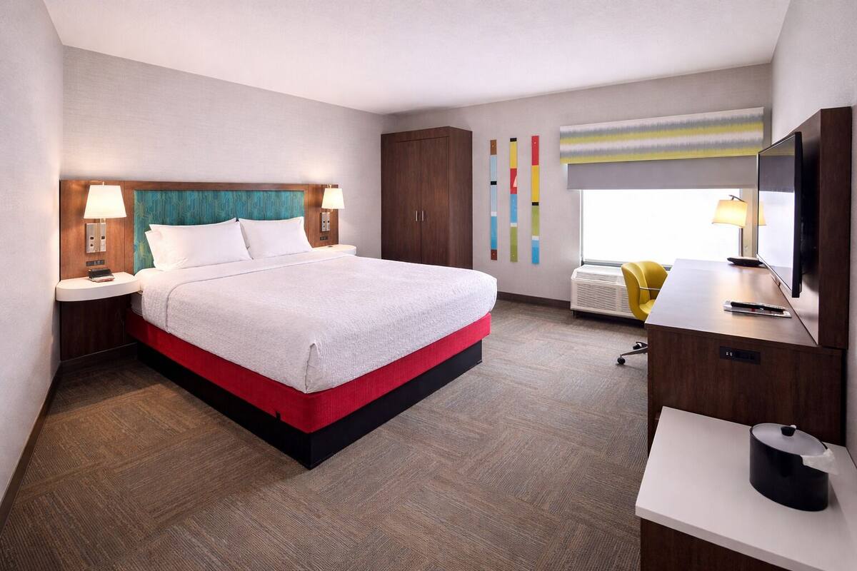 A hotel room with a bed, a desk, a TV, and a window with blinds.