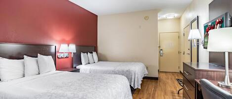 Habitación de hotel con dos camas, suelo de madera, pared de acento roja, escritorio con lámpara y puerta que da a un baño.