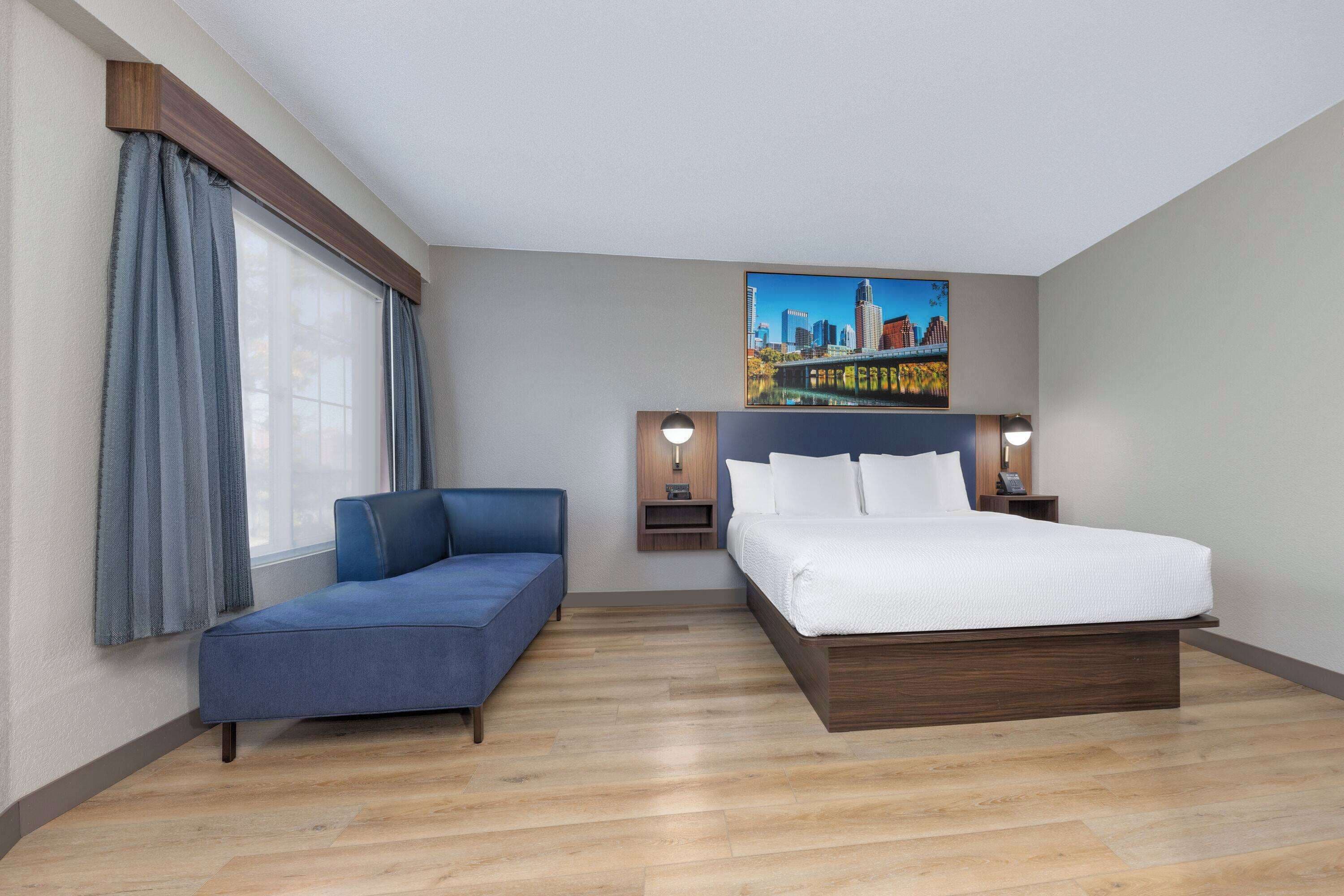 A hotel room with a bed, a blue sofa, a window with curtains, a cityscape painting, and a nightstand with a lamp.