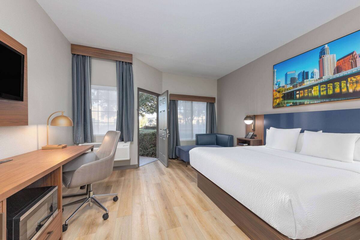 Habitación de hotel con una cama grande, un escritorio con silla, un televisor en la pared y un cuadro de un paisaje urbano.