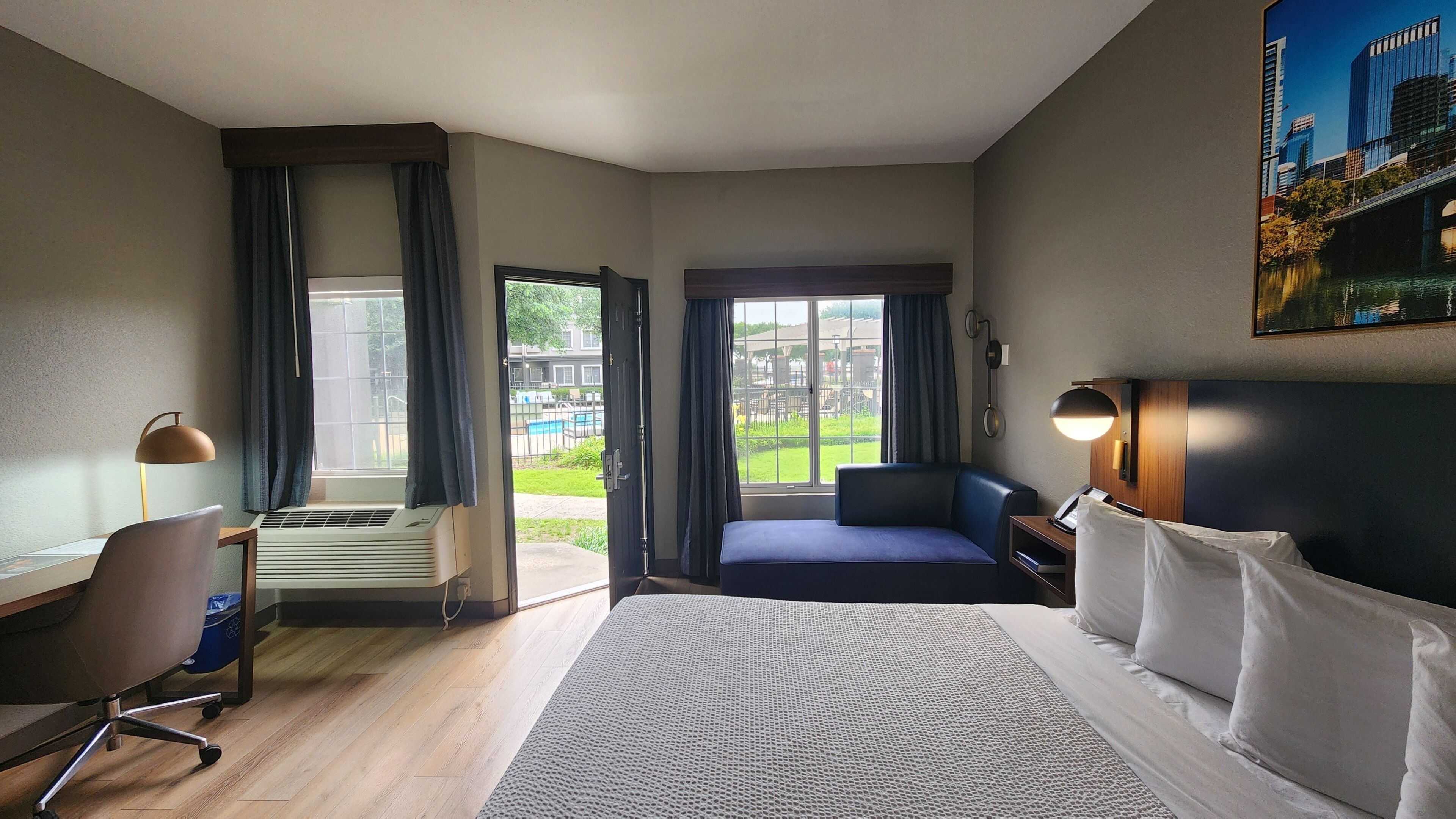 A hotel room with a bed, a desk, a chair, a window, and a view of a swimming pool and buildings.