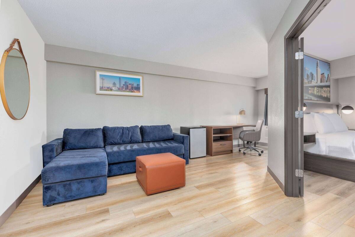 Una habitación de hotel moderna con un sofá modular, un pequeño taburete naranja, un escritorio con silla y una cama con vistas a la ciudad.