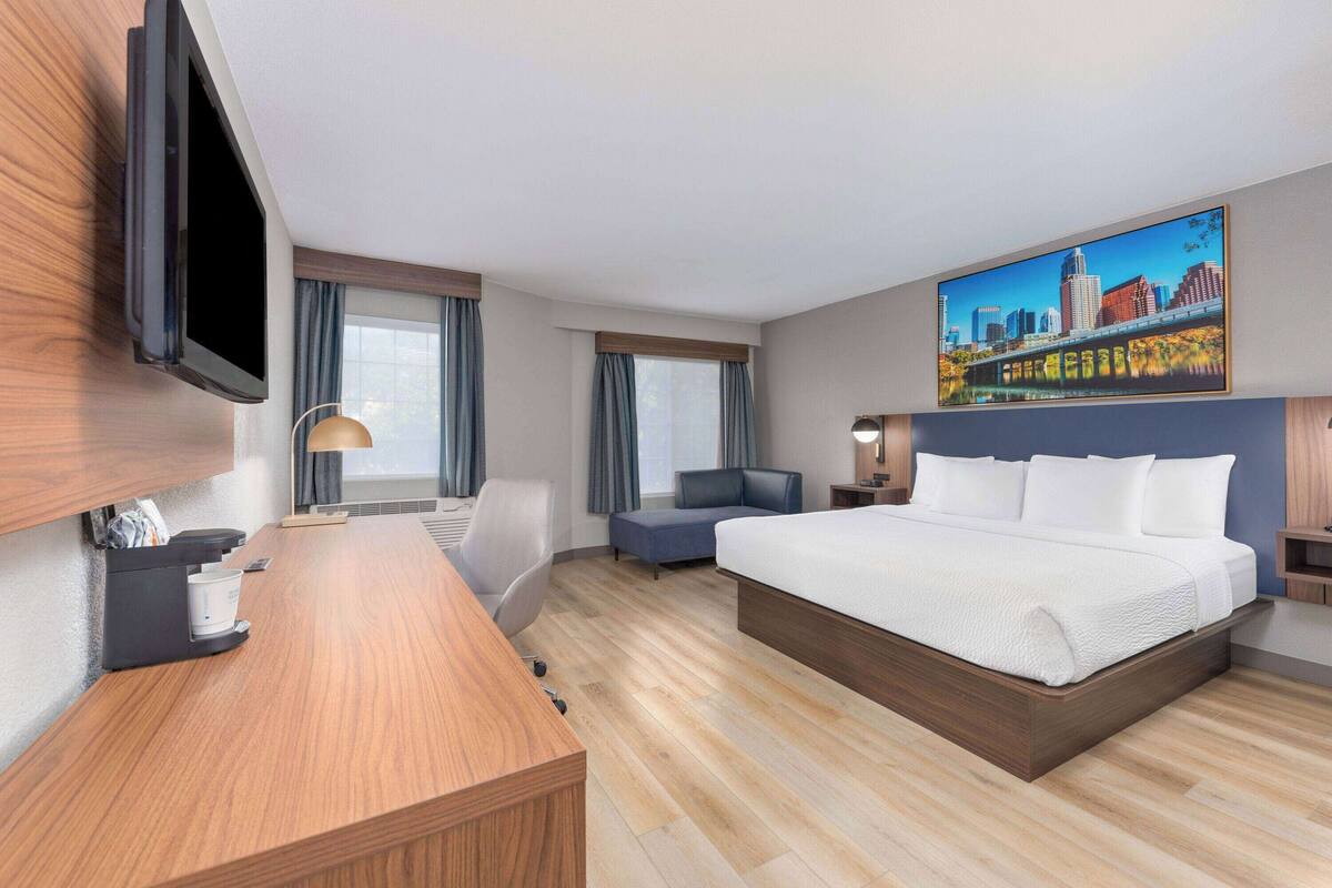 Habitación de hotel con una cama grande, un escritorio, un televisor de pantalla plana, un cuadro de un paisaje urbano y una ventana con cortinas.