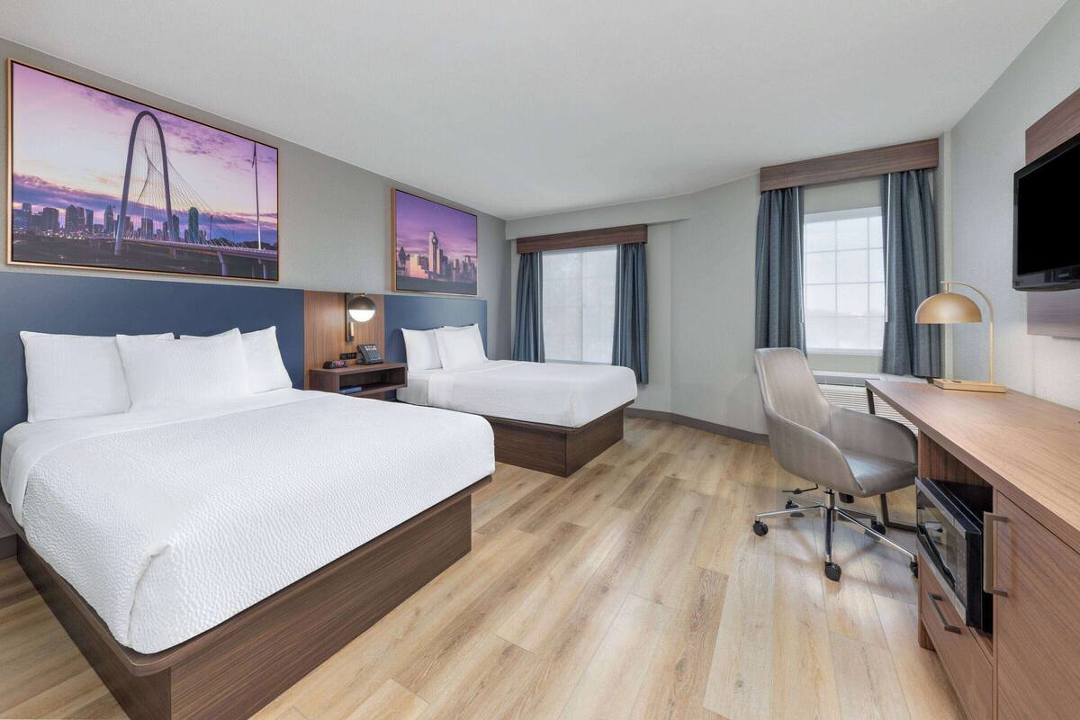 Habitación de hotel con dos camas, un escritorio, una silla, un televisor y ventanas grandes con cortinas.