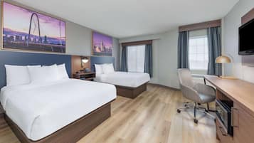 Habitación de hotel con dos camas, un escritorio, una silla, un televisor y ventanas grandes con cortinas.
