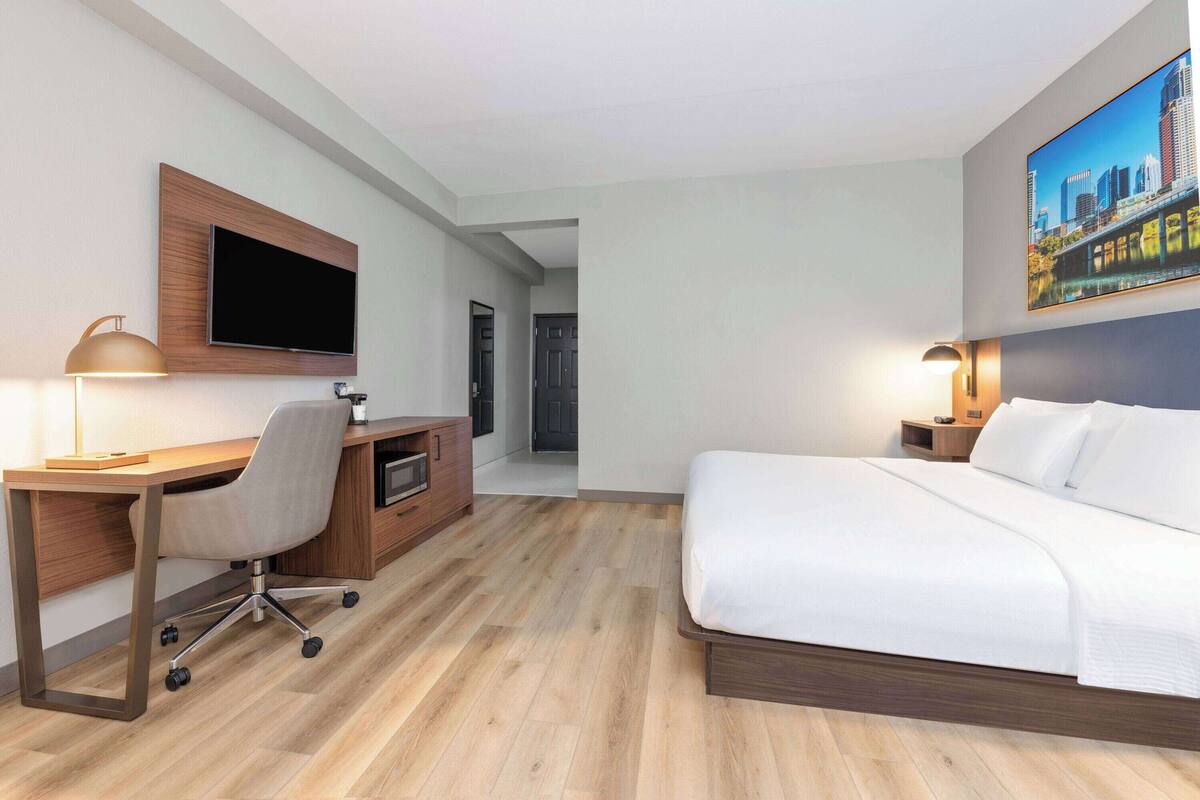 Una habitación de hotel con cama, escritorio, silla, televisor y un cuadro.