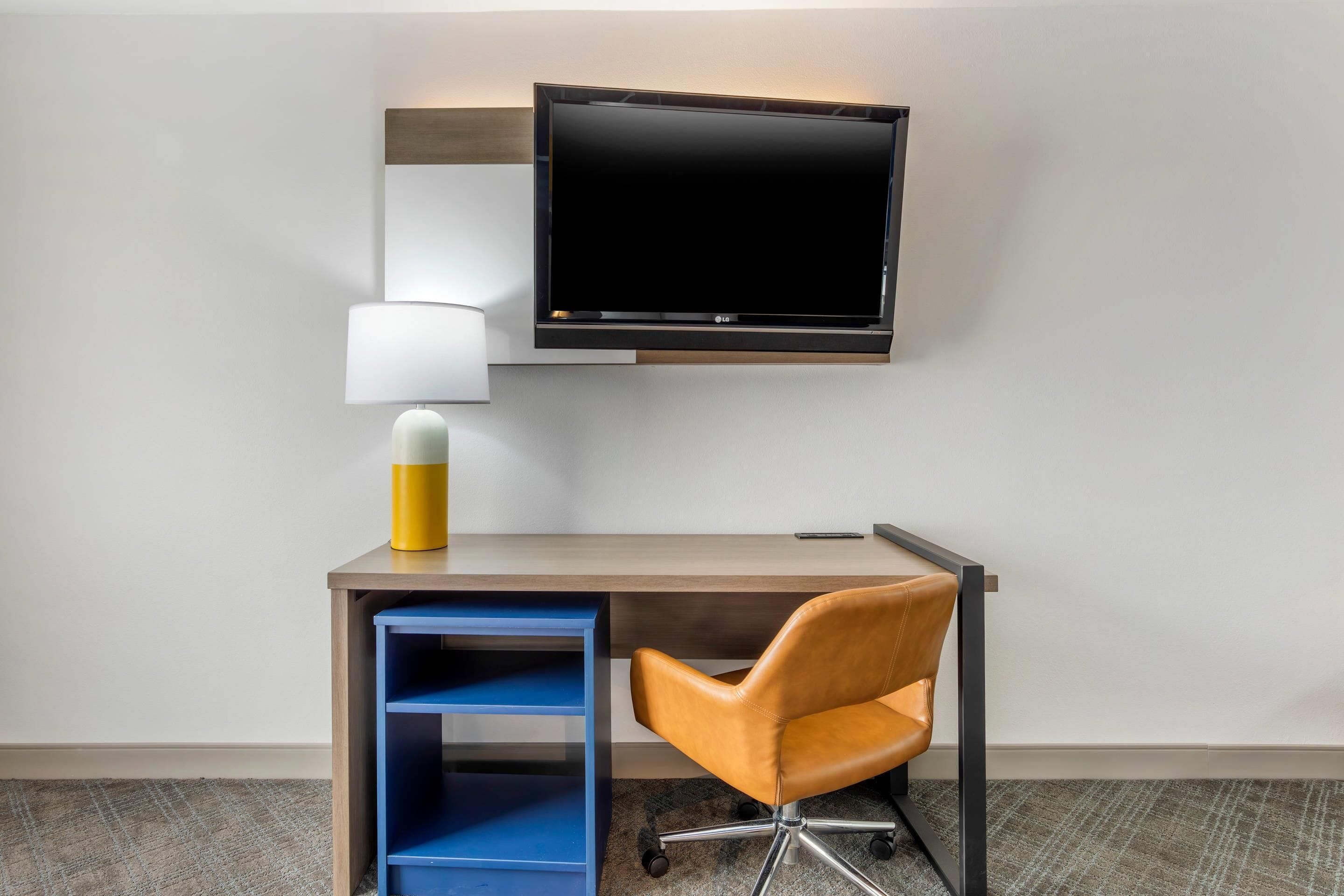 A hotel workspace with a desk, chair, TV, lamp, and storage unit.