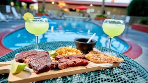 A dining table with a meal consisting of grilled meat, rice, and a plantain, accompanied by two drinks with lime garnishes, set against a poolside backdrop. - Family Garden Inn & Suites (Laredo)