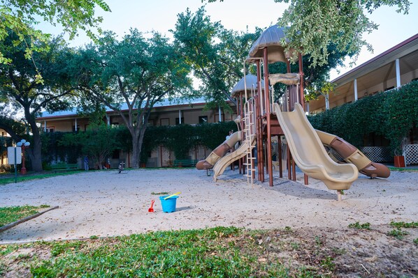 A playground with slides and climbing structures, surrounded by trees and a building in the background.