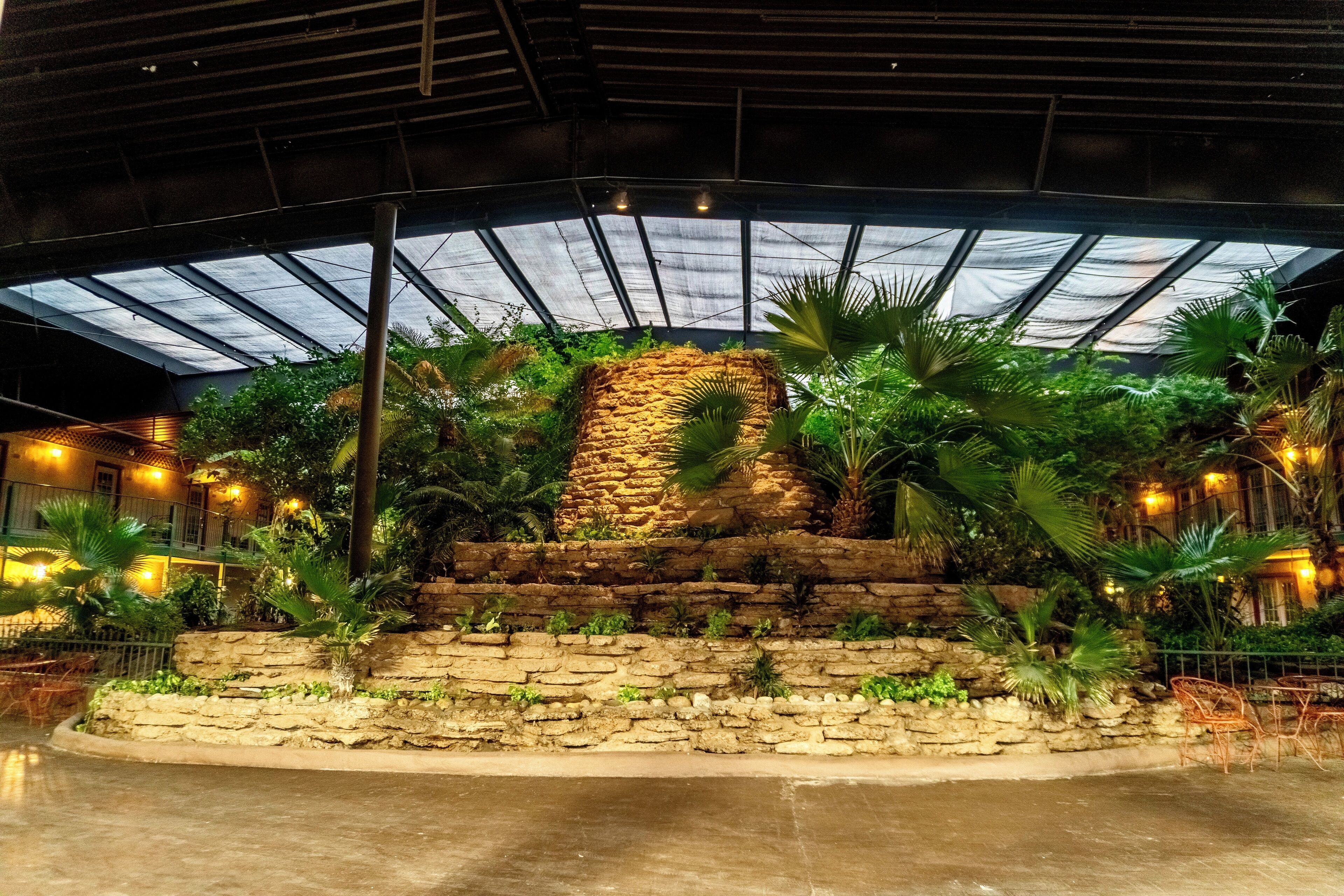 An indoor garden with a stone wall centerpiece, surrounded by tropical plants and furnished seating areas.