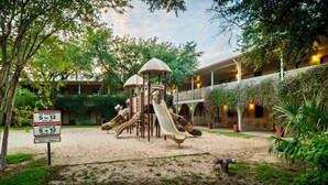 A playground with slides and a climbing structure, surrounded by greenery and a building with a balcony. - Family Garden Inn & Suites (Laredo)