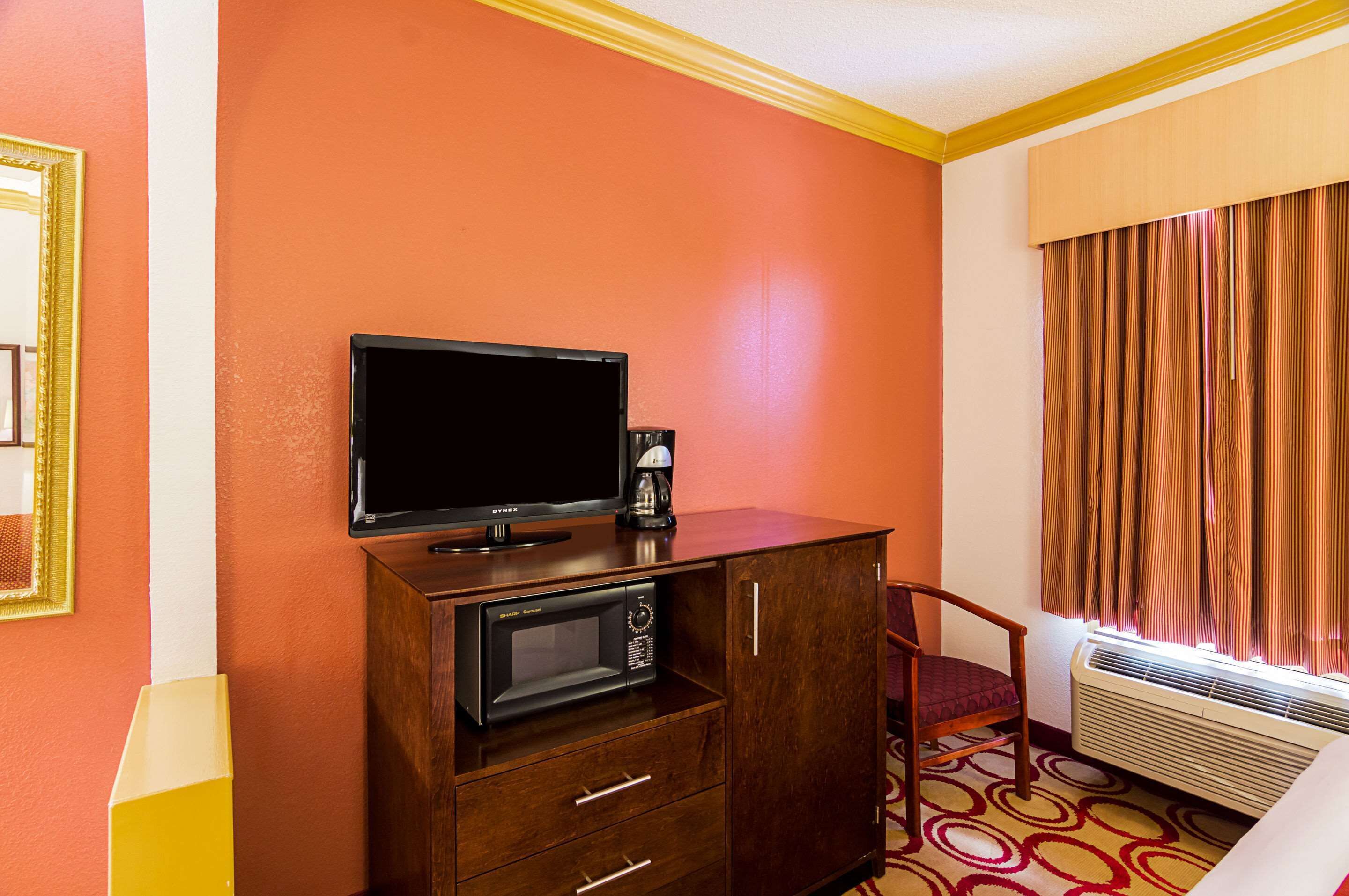 Een hotelkamer met een houten kastje waarin een televisie, een koffiezetapparaat en een magnetron zitten. Er is een stoel en een raam met gordijnen.