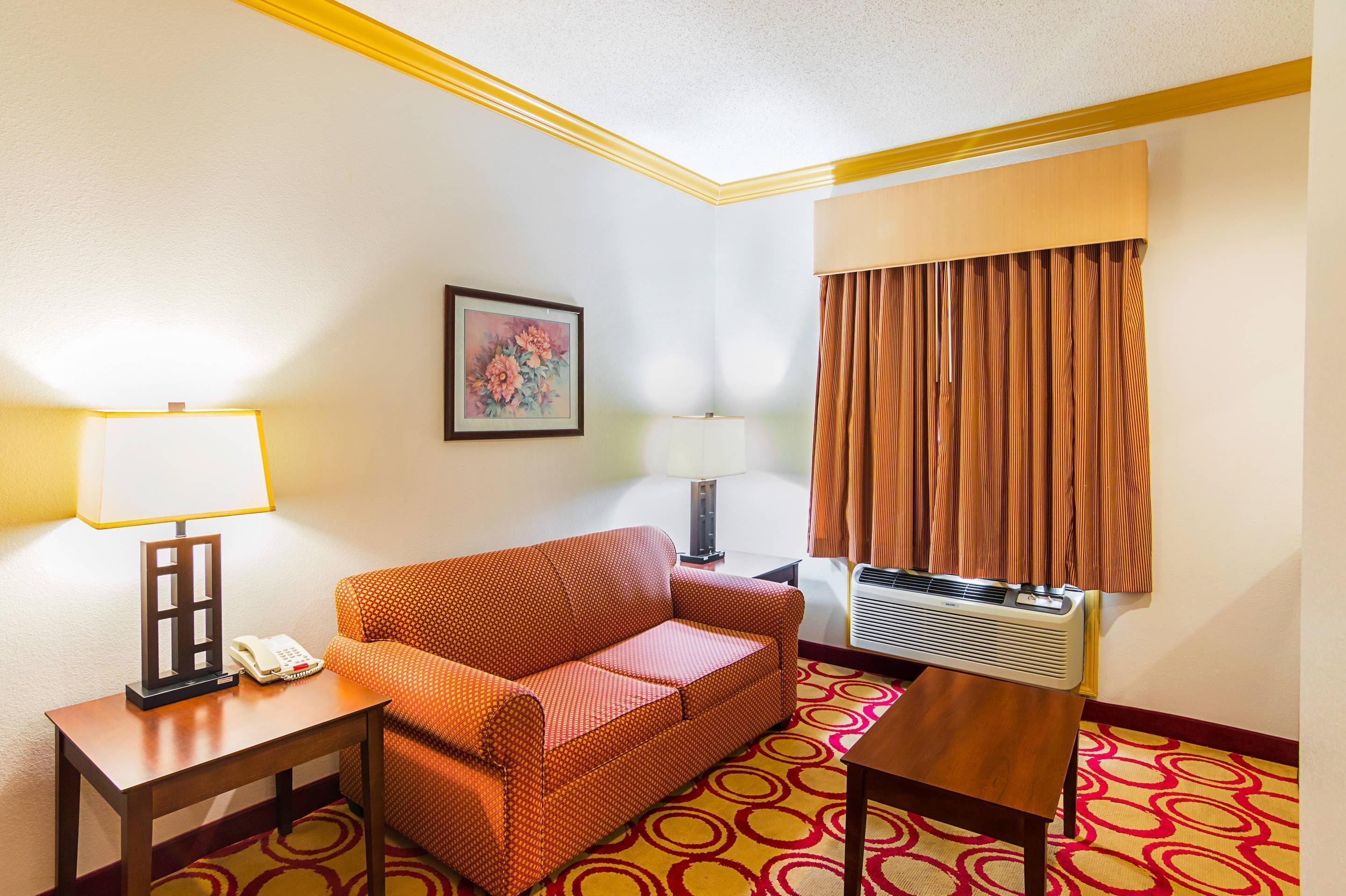 Une chambre d’hôtel avec un canapé rouge, deux tables de chevet avec des lampes, un climatiseur fixé au mur et une fenêtre avec des rideaux marron.