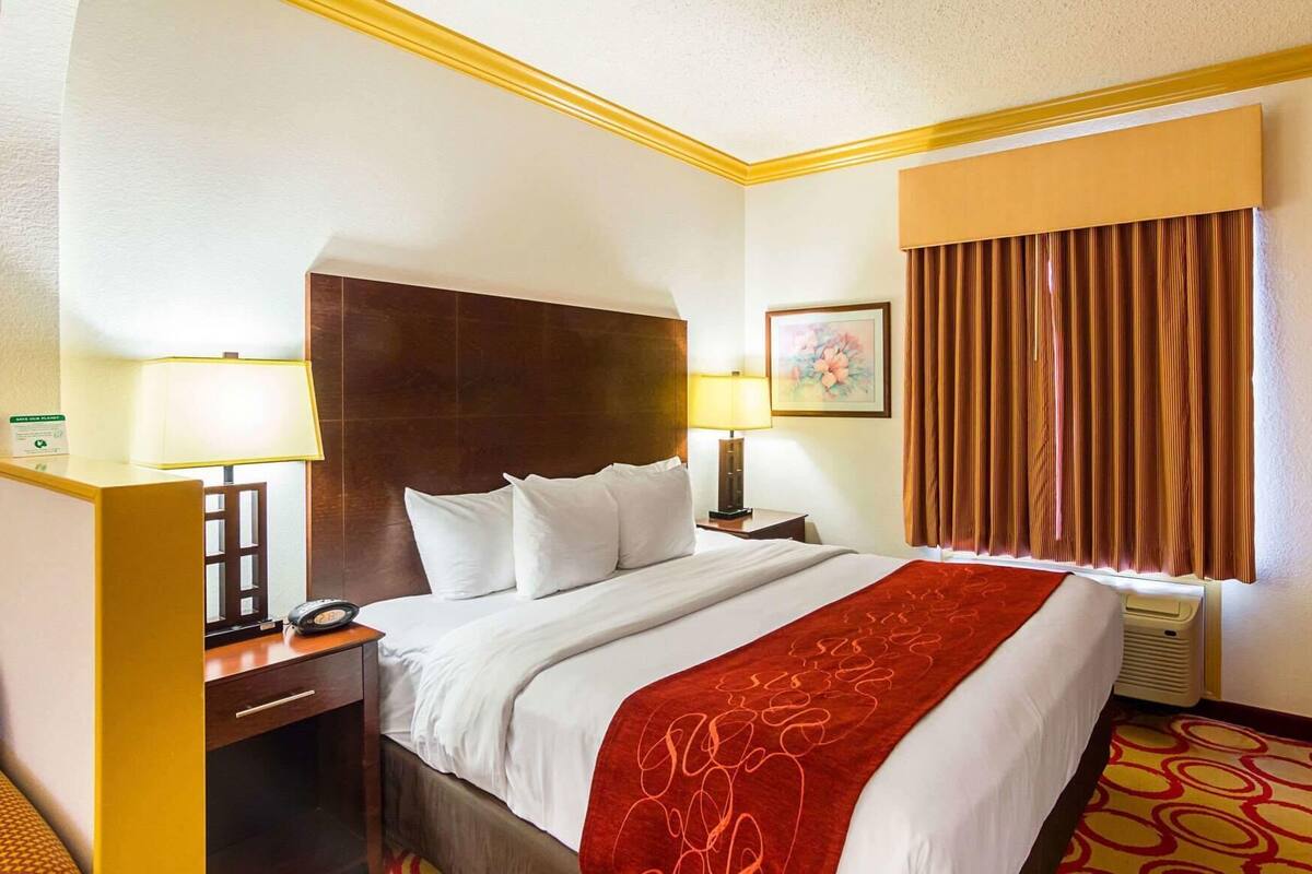A hotel room with a bed, two bedside tables, a nightstand, a lamp, a painting, and a window with curtains.