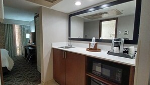 Une chambre d’hôtel équipée d’une kitchenette, comprenant un évier, une machine à café et un four à micro-ondes. Un miroir se trouve au-dessus de l’évier et une fenêtre est agrémentée de rideaux.