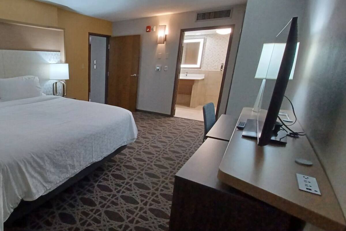 A hotel room with a large bed, a desk with a computer, a mirror, and a bathroom visible through an open door.