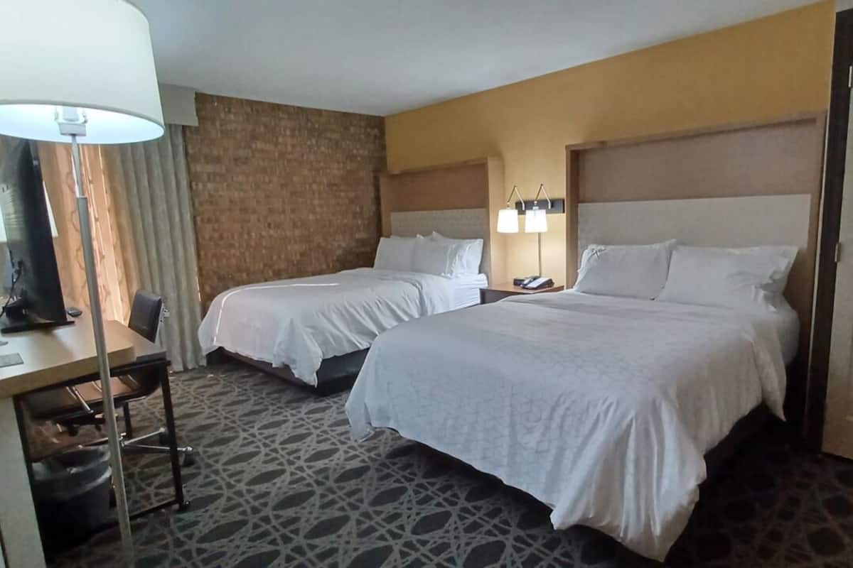 A hotel room with two beds, a desk with a lamp, a chair, a wardrobe, and a patterned carpet.
