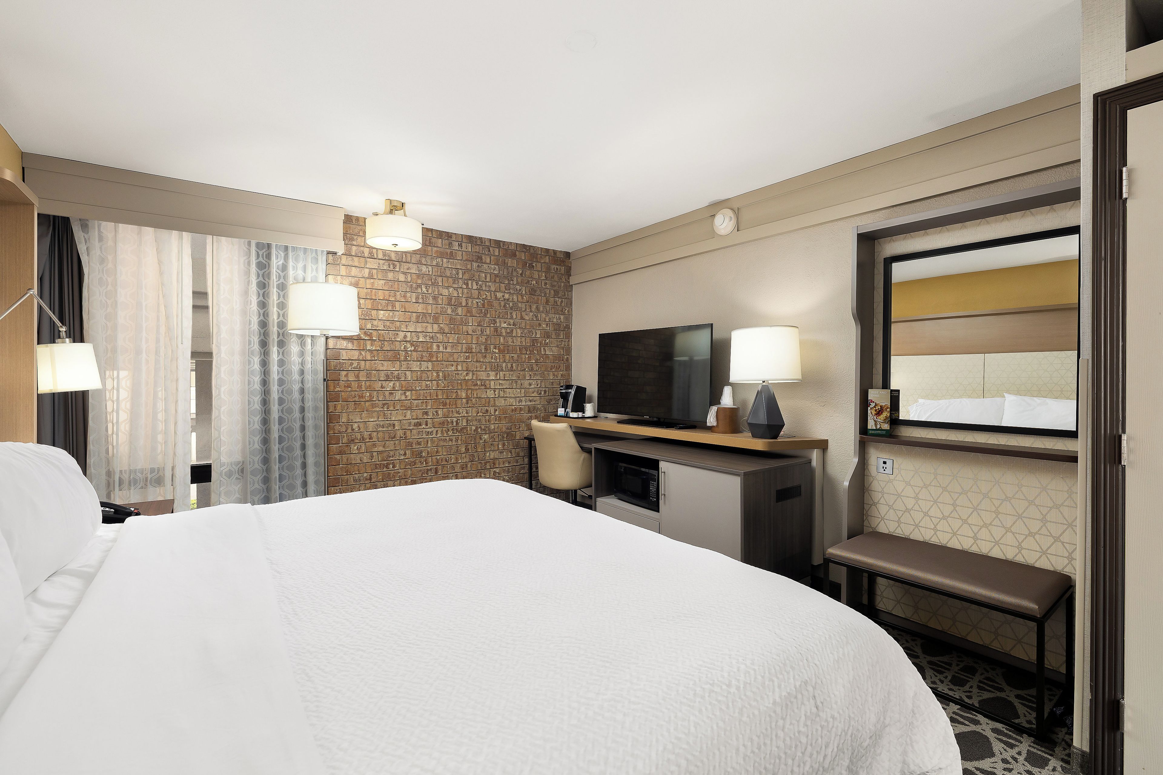 Ein Hotelzimmer mit einem großen Bett, einem Schreibtisch, einem Flachbildfernseher, einer Mikrowelle und einer Bank.